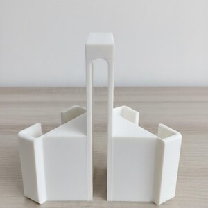 May include: Two white bookends with a minimalist design. Each bookend has a rectangular base and a triangular top, connected by a vertical support with an arched cutout. The bookends are made of a solid, opaque material.