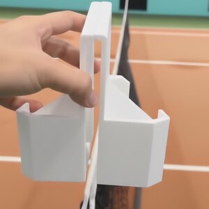 May include: A white plastic tennis net height gauge. The gauge is held in place by a hand and is positioned over a tennis net. The background shows a tennis court and foliage.