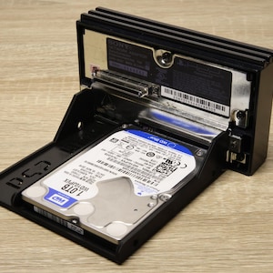 Puede incluir: Una carcasa de disco duro negra con un disco duro de 1,0 TB instalado. El disco duro está etiquetado como "WD Blue" y es parcialmente visible dentro de la carcasa abierta. La carcasa está abierta, revelando los componentes internos.