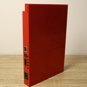 Peut inclure: Une console de jeux vidéo rouge vif positionnée verticalement. Le panneau latéral présente un port Ethernet métallique, un port de données rectangulaire, une série de fentes de ventilation verticales et une prise d'alimentation à deux broches.