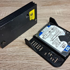 May include: A black plastic hard drive adapter for a game console, featuring a 1.0TB WD hard drive mounted in a bracket with the text PS2 embossed on the side, alongside a matching black plastic cover with a yellow warning sticker attached.