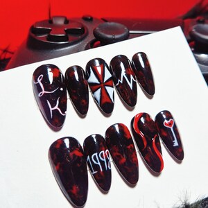 Pode incluir: Um conjunto de dez unhas artificiais longas e ovais. Cada unha é pintada com um design marmorizado em vermelho escuro e preto. Algumas unhas apresentam gráficos brancos, incluindo as letras "L K", uma cruz branca e vermelha, uma linha de batimento cardíaco, o número "1998", um redemoinho vermelho e uma chave com um coração.