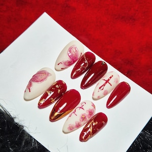 Pode incluir: Um conjunto de dez unhas postiças em forma de amêndoa. As unhas apresentam uma variedade de designs, incluindo vermelho liso, detalhes dourados, padrões florais e um design de cruz. A paleta de cores inclui vermelho, branco e dourado.