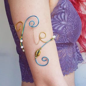 Op de afbeelding: Een bovenarmband gemaakt van blauwe en goudkleurige draad in krullende patronen. Het sieraad is versierd met kleine witte kralen en een enkele goudkleurige kaurischelp, verbonden door een fijn goudkleurig metalen kettinkje.