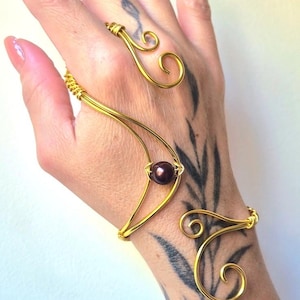 Pode incluir: Um ornamento de mão em fio metálico dourado, composto por um anel ligado a uma pulseira por uma estrutura de fio curvo. Uma única conta esférica castanho-escura está centrada na estrutura de fio, decorada com extremidades em espiral.