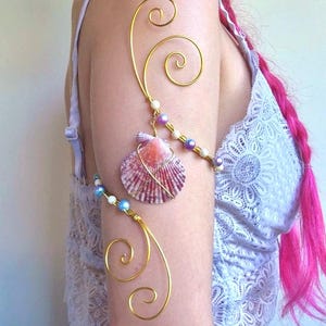 Pode incluir: Uma braçadeira de fio dourado com padrões em espiral, decorada com pequenas contas brancas e roxas iridescentes. Uma única concha de vieira rosa e branca está suspensa no fio, presa com um fino enrolamento de fio metálico dourado.