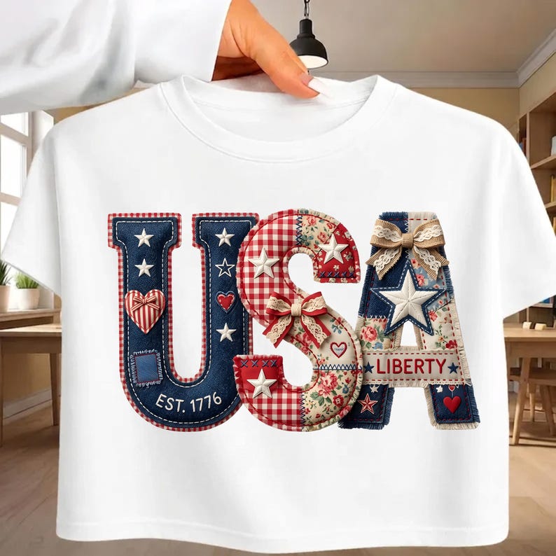May include: A white t-shirt featuring a large graphic of the letters USA. The U is dark blue with white stars and a heart. The S is red and white gingham with floral patterns and bows. The A features a star, a bow, and the text LIBERTY.
