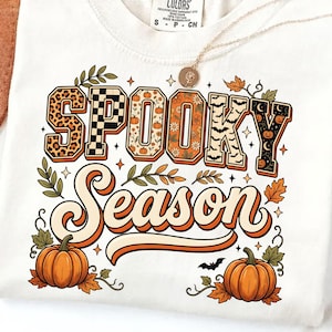 Puede incluir: Una camiseta blanca con el texto SPOOKY SEASON en letras de bloque y cursiva. Las letras están rellenas con patrones que incluyen estampado de leopardo, cuadros, calabazas, flores, murciélagos y lunas. Dos calabazas naranjas y hojas de otoño rodean el texto.