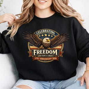 Può includere: Felpa nera con un design patriottico con un'aquila. Il design include le parole "Celebrating Freedom" e "250 Years of Liberty 1776-Anniversary-2026". L'aquila è raffigurata con le ali spiegate, su uno sfondo circolare.