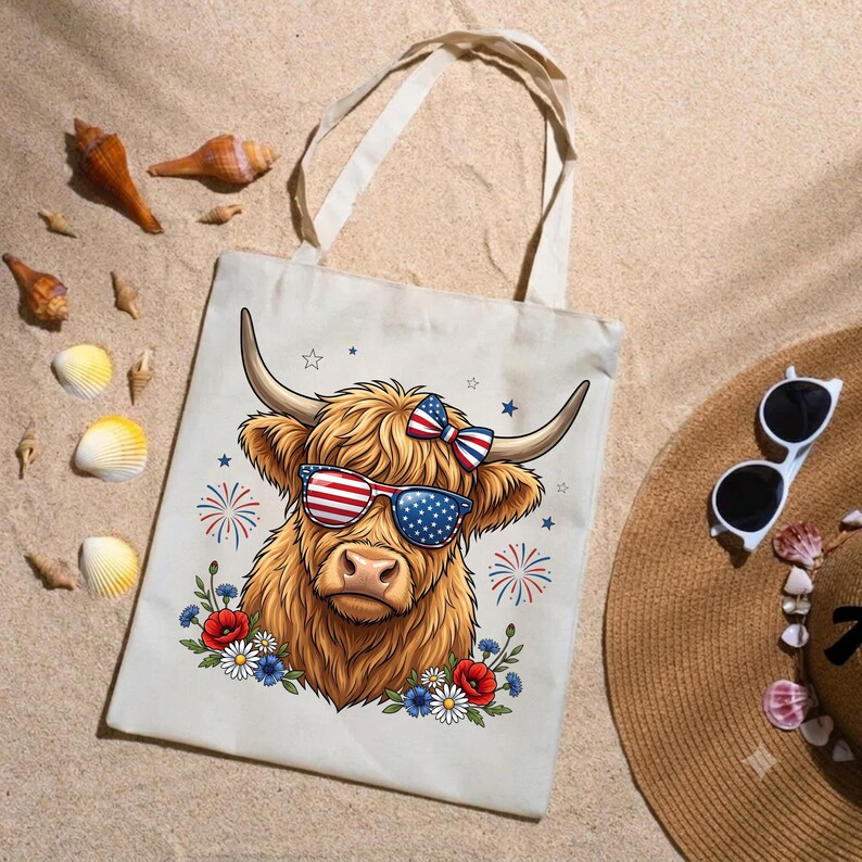 May include: A cream-colored canvas tote bag featuring a graphic of a Highland cow wearing American flag-patterned sunglasses and a matching bow. The design includes red, white, and blue fireworks, stars, and a floral arrangement of poppies and daisies.
