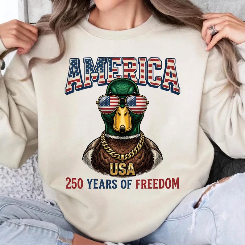 May include: A cream-colored sweatshirt featuring a graphic of a mallard duck wearing American flag sunglasses and a gold chain. The text reads AMERICA above the duck, and USA 250 YEARS OF FREEDOM below the duck in red and blue lettering.