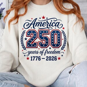 May include: A cream-colored crewneck sweatshirt featuring a graphic design with the text America 250 years of freedom 1776-2026. The design includes large blue numbers filled with white stars, red and blue outlines, and red star accents.