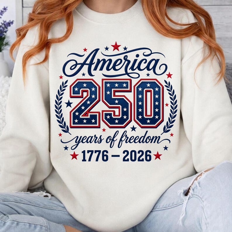 May include: A cream-colored crewneck sweatshirt featuring a graphic design with the text America 250 years of freedom 1776-2026. The design includes large blue numbers filled with white stars, red and blue outlines, and red star accents.