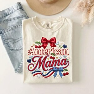 May include: A cream-colored t-shirt featuring a graphic print with the text American Mama in red script. The design includes a large red bow, red cherries, blue hearts, small white flowers, gold stars, and a red, white, and blue ribbon accent.