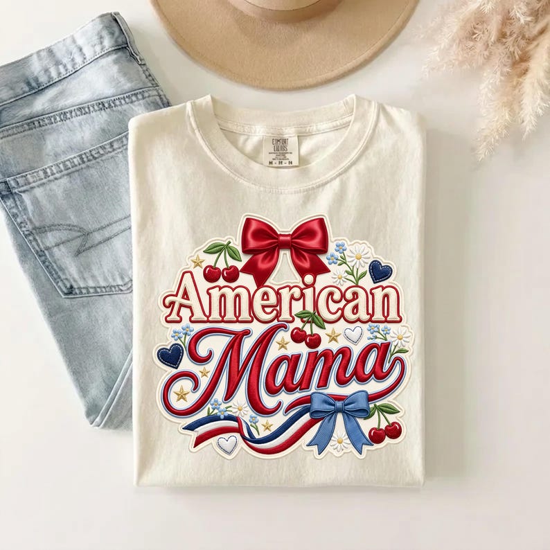 May include: A cream-colored t-shirt featuring a graphic print with the text American Mama in red script. The design includes a large red bow, red cherries, blue hearts, small white flowers, gold stars, and a red, white, and blue ribbon accent.