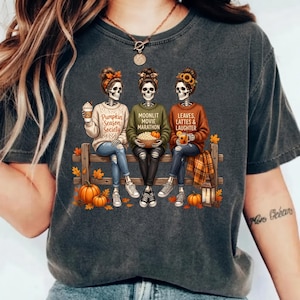 May include: A charcoal grey t-shirt featuring a graphic of three skeletons sitting on a wooden fence. The skeletons wear jumpers with the text Pumpkin Season Society, Moonlit Movie Marathon, and Leaves, Lattes & Laughter, surrounded by pumpkins.