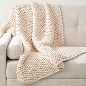 May include: A cream-colored, chunky knit throw blanket draped over a light-colored sofa. The blanket has a textured, raised stitch pattern and is folded to display its thickness and warmth. The sofa has a neutral color and a simple design.