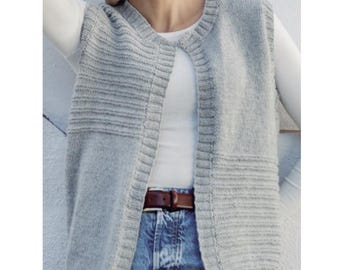 May include: A light gray sleeveless knit vest featuring a ribbed crew neckline, a vertical ribbed front placket, and horizontal ribbed panels on the upper chest and lower sides. The garment is worn over a white long-sleeve shirt and blue jeans.