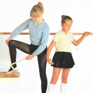May include: Two young ballet dancers in a studio. One wears a light blue wrap sweater, black tights, and pointe shoes. The other wears a white V-neck shirt, a black skirt, and white knee socks. A wooden barre is in the background.