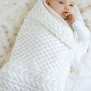 May include: A baby wrapped in a white knitted blanket with cable knit and textured patterns. The blanket is soft and cozy, perfect for swaddling. The baby is wearing a white long-sleeved shirt.