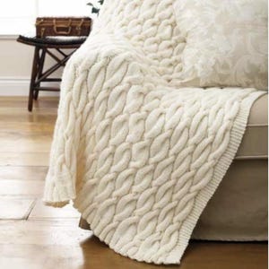 May include: A cream-colored knitted throw blanket featuring a prominent cable knit pattern with vertical braided textures. The blanket is draped over a piece of furniture, showcasing a thick, soft yarn construction with a finished ribbed edge border.