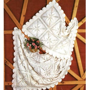 May include: A white crocheted blanket featuring a repeating floral pattern with intricate openwork details and a scalloped edge. A small woven basket containing a cluster of pink and green artificial flowers rests on the fabric surface.