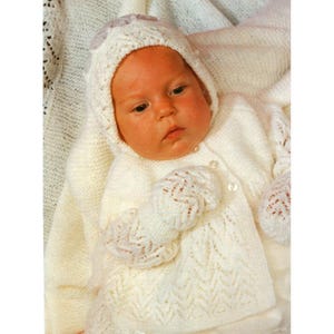 May include: A baby wearing a white knitted outfit, including a hooded sweater, mittens, and a matching hat. The outfit has a delicate, openwork pattern and is buttoned in the front. The baby is lying on a soft, white blanket.