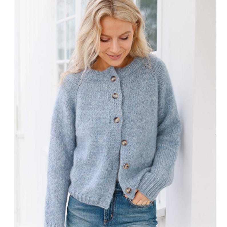 May include: A light blue cardigan sweater with a round neckline and button-up front. The sweater has long sleeves and is made of a soft, textured knit fabric. The buttons are a light brown color. The person is wearing blue jeans.