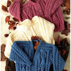 Könnte beinhalten: Drei Zopfstrick-Cardigans in Rosa-, Creme- und Blautönen. Die Pullover haben Knopfverschlüsse und aufwendige Webmuster. Herbstblätter und Kastanien sind um die Kleidungsstücke herum verstreut.