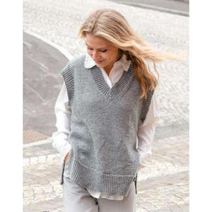 May include: A gray knitted V-neck vest with ribbed detailing. The vest is worn over a white collared shirt and light-colored pants. The model is outside on a paved surface.