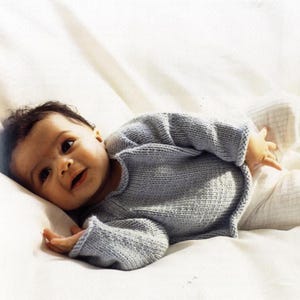 May include: A baby wearing a light blue knitted sweater and white pants lies on a white surface. The sweater has long sleeves and a rounded neckline. The baby is looking at the camera.