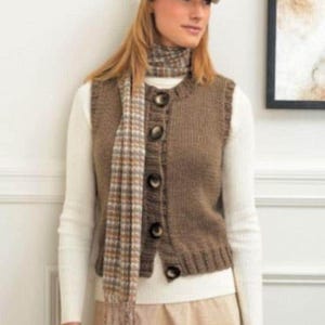 Può includere: Un gilet marrone lavorato a maglia con grandi bottoni scuri. Il gilet è indossato sopra una camicia bianca a maniche lunghe e abbinato a una sciarpa abbinata. La sciarpa ha un motivo a quadri nei toni del marrone, beige e bianco. Un berretto completa l'outfit.