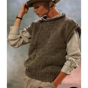 Könnte beinhalten: Eine braune Strickweste mit vertikalem Korbflechtmuster auf der Vorderseite, getragen über einem langärmeligen Hemd mit kleinem weiß-braunem Karomuster. Das Outfit umfasst einen breitkrempigen Hut und eine hellfarbene Hose.