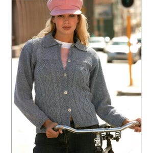May include: A gray cable-knit cardigan with a collared neckline and button-up front. The long-sleeved sweater features a textured pattern and is worn with a pink cap. The person is holding bicycle handlebars.