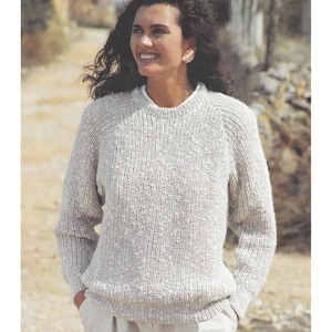 May include: A light grey knitted sweater with a crew neck and long sleeves. The sweater has a textured appearance and a relaxed fit. The model is wearing the sweater with a white undershirt and light-coloured trousers. The background is a blurred outdoor scene.