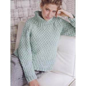 May include: A light green, textured knit sweater with a high, ribbed collar and long sleeves. The sweater has a subtle, all-over pattern and is made of a soft, comfortable material. The model is sitting on a white couch.