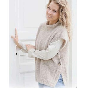 Op de afbeelding: Een beige mouwloze trui met een hoge halslijn en twee knopen aan de zijkant. De trui wordt over een wit overhemd met kraag gedragen. De vrouw op de foto lacht en leunt tegen een witte deur.