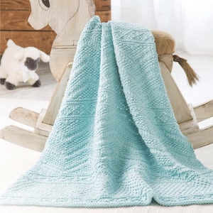 May include: A light turquoise knitted blanket featuring a textured diamond pattern with alternating sections of seed stitch and smooth stockinette. The fabric has a consistent soft color throughout and is draped over a wooden rocking horse structure.