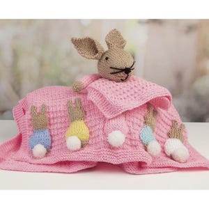 May include: A pink knitted security blanket featuring a stuffed rabbit head with long ears and black embroidered whiskers. The blanket is decorated with four small knitted rabbit figures in blue, yellow, light blue, and white outfits.