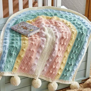 May include: A semicircular crochet blanket featuring concentric stripes in pastel blue, mint green, yellow, pink, and cream. The texture includes raised bobble stitches throughout the design. Four large cream-colored yarn pom-poms hang from the bottom edge.