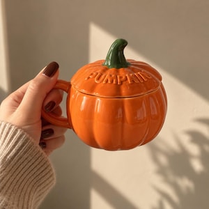 Könnte beinhalten: Ein glänzender, orangefarbener Keramikbecher in Kürbisform mit passendem Deckel. Der Deckel hat einen grünen Stiel als Griff und das Wort PUMPKIN ist in Großbuchstaben eingeprägt. Der Becher verfügt über einen geschwungenen Henkel an der Seite.