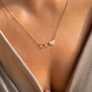May include: A delicate gold necklace featuring three heart pendants. The first two hearts are open, while the third heart has the letter "A" engraved on it. The necklace is displayed on a person's chest, against a neutral background.