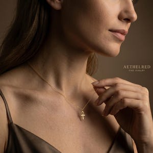 May include: A gold-colored ankh pendant necklace. The ankh is suspended from a delicate chain. The image is set against a soft, neutral background. The text "AETHELRED FINE JEWELRY" is visible in the upper right corner.