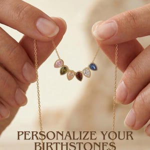 Könnte beinhalten: Eine goldene Halskette mit einer Kette und einer Reihe von tropfenförmigen Edelsteinen in verschiedenen Farben. Die Halskette wird von Händen gehalten, und der Text "PERSONALIZE YOUR BIRTHSTONES" wird darunter angezeigt. Die Edelsteine sind rosa, grün, pfirsichfarben, rot, weiß, blau.