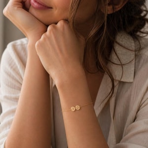 Puede incluir: Una delicada pulsera dorada con dos pequeños colgantes redondos con iniciales grabadas. La pulsera se lleva en la muñeca, centrándose en la joya. El fondo está suavemente borroso, resaltando los detalles de la pulsera.