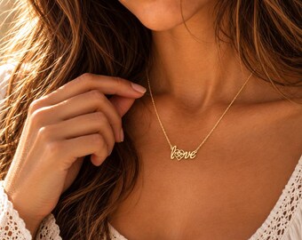 Pode incluir: Um colar de tom dourado com uma corrente fina e um pendente com a palavra love, onde a letra o é substituída por um pequeno coração com um design floral recortado no seu interior.