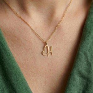 May include: A gold-colored necklace with a delicate chain. The pendant features the letter "H" with a heart shape incorporated into the design. The necklace is displayed against a green background.