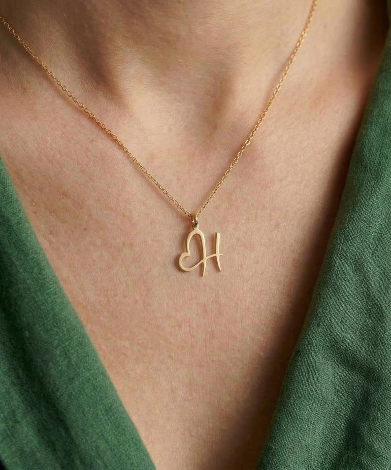 May include: A gold-colored necklace with a delicate chain. The pendant features the letter "H" with a heart shape incorporated into the design. The necklace is displayed against a green background.