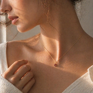 May include: A delicate gold necklace with a small, teardrop-shaped, red gemstone pendant. The necklace is worn on a person's neck, and the gold chain is thin and elegant. The pendant is a vibrant red color.
