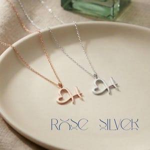 May include: Two delicate necklaces with heart-shaped pendants and the letter "H". One is rose gold, the other silver. The necklaces are displayed on a white plate. The text "ROSE SILVER" is below the necklaces.
